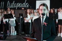 Na zdjęciu widoczny jest apel z okazji Dnia Niepodległości, obchodzonego 11 listopada. Na pierwszym planie stoi uczennica, ubrana elegancko w czarną marynarkę, z przypiętą biało-czerwoną kokardką, symbolizującą polski patriotyzm. Trzyma mikrofon oraz notatnik. W tle znajduje się grupa uczniów, ubranych w galowe stroje, przygotowujących się do występu.