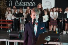 Na zdjęciu widoczny jest apel z okazji Dnia Niepodległości, obchodzonego 11 listopada. Na pierwszym planie stoi uczeń, ubrany elegancko w czarną marynarkę, z przypiętą biało-czerwoną kokardką, symbolizującą polski patriotyzm. Trzyma mikrofon oraz notatnik. W tle znajduje się grupa uczniów, ubranych w galowe stroje, przygotowujących się do występu.