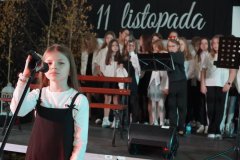 Na pierwszym planie widać uczennicę przy mikrofonie, przygotowującą się do wystąpienia. Jest ubrana w biały podkoszulek i czarną sukienkę na ramiączkach. W tle stoi grupa uczniów w odświętnych strojach, gotowych do wykonania części artystycznej. 