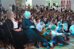  Na zdjęciu widzimy apel szkolny z okazji Dnia Niepodległości. W pierwszym planie siedzą dyrektorki szkoły, ubrane odświętnie, z przypiętymi biało-czerwonymi kokardami symbolizującymi patriotyzm. Za nimi widać dużą grupę uczniów ubranych na biało, siedzących na podłodze oraz stojących w tle. Cała sala wypełniona jest uczniami i nauczycielami, którzy biorą udział w uroczystościach. W tle widać polskie flagi oraz osoby obserwujące wydarzenie.