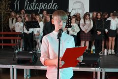  Na zdjęciu widoczny jest apel z okazji Dnia Niepodległości, obchodzonego 11 listopada. Na pierwszym planie stoi uczeń, ubrany elegancko, z przypiętą biało-czerwoną kokardką, symbolizującą polski patriotyzm. Trzyma mikrofon oraz notatnik. 