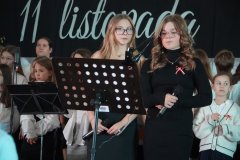  Na pierwszym planie widać dwie uczennice przy mikrofonie, przygotowującą się do wystąpienia. W tle stoi grupa uczniów w odświętnych strojach, gotowych do wykonania części artystycznej. 