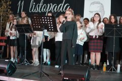  Na pierwszym planie widać  uczennicę przy mikrofonie, przygotowującą się do wystąpienia. W tle stoi grupa uczniów w odświętnych strojach, gotowych do wykonania części artystycznej. 