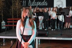  Na pierwszym planie widać  uczennicę przy mikrofonie, przygotowującą się do wystąpienia. W tle stoi grupa uczniów w odświętnych strojach, gotowych do wykonania części artystycznej. 