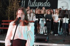 Na pierwszym planie widać  uczennicę przy mikrofonie, przygotowującą się do wystąpienia. W tle stoi grupa uczniów w odświętnych strojach, gotowych do wykonania części artystycznej. 