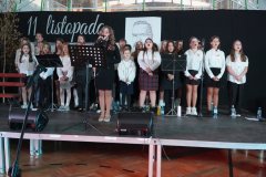  Na zdjęciu widać szkolną scenę podczas apelu z okazji Dnia Niepodległości. Na scenie znajduje się grupa uczniów ubranych w odświętne stroje, śpiewających patriotyczne pieśni. W tle widoczna jest dekoracja z napisem „11 listopada” oraz biało-czerwona flaga. Obok grupy stoi solistka przy mikrofonie. 
