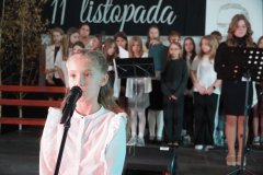  Na pierwszym planie widać  uczennicę przy mikrofonie, przygotowującą się do wystąpienia. W tle stoi grupa uczniów w odświętnych strojach, gotowych do wykonania części artystycznej. 