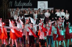 Na zdjęciu widać występ uczniów podczas apelu z okazji Dnia Niepodległości. W pierwszym planie dzieci w biało-czerwonych strojach wykonują układ taneczny z wykorzystaniem flag i wstążek w barwach narodowych, a dziewczynki mają na głowach wianki z kwiatów. W tle na scenie znajduje się chór uczniów, ubranych odświętnie, śpiewających patriotyczne pieśni. Na dekoracji widoczny jest napis „11 listopada” oraz portret wykonany z tekstu, upamiętniający ważną postać historyczną. Atmosfera jest podniosła i pełna patriotyzmu.
