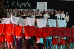 Na zdjęciu widać występ uczniów podczas apelu z okazji Dnia Niepodległości. W pierwszym planie dzieci w biało-czerwonych strojach wykonują układ taneczny z wykorzystaniem flag i wstążek w barwach narodowych, a dziewczynki mają na głowach wianki z kwiatów. W tle na scenie znajduje się chór uczniów, ubranych odświętnie, śpiewających patriotyczne pieśni. Na dekoracji widoczny jest napis „11 listopada” oraz portret wykonany z tekstu, upamiętniający ważną postać historyczną. Atmosfera jest podniosła i pełna patriotyzmu.