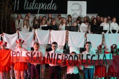 Na zdjęciu widać występ uczniów podczas apelu z okazji Dnia Niepodległości. W pierwszym planie dzieci w biało-czerwonych strojach wykonują układ taneczny z wykorzystaniem flag i wstążek w barwach narodowych, a dziewczynki mają na głowach wianki z kwiatów. W tle na scenie znajduje się chór uczniów, ubranych odświętnie, śpiewających patriotyczne pieśni. Na dekoracji widoczny jest napis „11 listopada” oraz portret wykonany z tekstu, upamiętniający ważną postać historyczną. Atmosfera jest podniosła i pełna patriotyzmu.