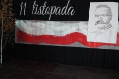 Na zdjęciu przedstawiona jest dekoracja z okazji apelu związanego z obchodami Święta Niepodległości 11 listopada. W centralnej części widoczna jest biało-czerwona flaga Polski wykonana z pociągnięć farby na tle czarnego materiału. Nad flagą umieszczono napis „11 listopada”. Po prawej stronie znajduje się portret Józefa Piłsudskiego, utworzony z drobnych napisów, co przypomina technikę typograficzną. Dekoracja jest utrzymana w patriotycznej tonacji, podkreślając symbolikę polskiej niepodległości i oddając hołd bohaterom narodowym.