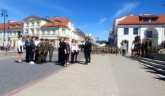Grupa młodych ludzi i młodzieży ustawiona w rzędzie na placu miejskim; część w mundurach wojskowych, część w ubraniach cywilnych. Naprzeciw nich stoi oficer lub instruktor. W tle rząd żołnierzy i zabudowa miejska z budynkami o czerwonych dachach, przy słonecznym, bezchmurnym niebie.