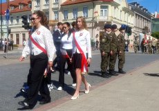 Na zdjęciu widać pochód upamiętniający święto narodowe. W pierwszym planie idą młode osoby — prawdopodobnie uczennice — ubrane w biało-czerwone szarfy i kokardy, co sugeruje barwy narodowe Polski. Kilka dziewcząt ma eleganckie stroje: białe bluzki i ciemne spódnice lub spodnie; jedna nosi białe szpilki. Za nimi po prawej maszeruje grupa młodych ludzi w mundurach wojskowych, ustawionych w dwóch szeregach.