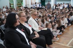 W sali gimnastycznej podczas szkolnej uroczystości: na pierwszym planie Dyrekcja Szkoły — ubrane oficjalnie, w jasnych i ciemnych marynarkach, z przypiętymi biało czerwonymi kotylionami; za nimi tłum dzieci siedzących na podłodze i stojących w rzędach, ubrane głównie w biało ciemne stroje.