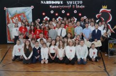 Grupowe zdjęcie szkolne na tle dekoracji upamiętniającej Święto Konstytucji 3 Maja: kilkadziesiąt dzieci i kilku dorosłych ustawionych w kilku rzędach; część uczniów ma przypięte biało czerwone kotyliony, niektórzy ubrani są w czerwone koszulki z motywem flagi, inni w białych koszulach i garniturach. Za nimi duża czarna ścianka z napisem „NARODOWE ŚWIĘTO Konstytucji 3 Maja” Zdjęcie  po szkolnej uroczystości.
