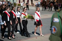 Na zdjęciu grupa młodych osób uczestniczy w ceremonii pamięci. Idą w rzędzie po wybrukowanym placu; kilka z nich nosi biało-czerwone szarfy przewieszone przez ramię, jedna niesie bukiet kwiatów, prawdopodobnie do złożenia pod pomnikiem. Ubrania są formalne — ciemne spodnie lub spódnice i białe bluzki/koszule.
