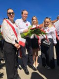 Grupa czterech osób stojących na zewnątrz na tle niebieskiego nieba i budynków; trzy kobiety i jeden mężczyzna ubrani w białe koszule i ciemne spodnie/spódnice, każda osoba ma na piersi kokardę w biało-czerwonych barwach, jedna kobieta trzyma małą flagę Polski, mężczyzna oraz kobieta trzymają razem bukiet czerwono-białych kwiatów.