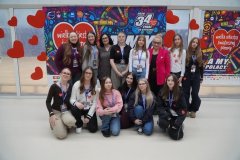 Zdjęcie przedstawia  grupę dwunastu dziewcząt, wolontariuszek WOŚP  wraz z Dyrekcją Szkoły Panią Marleną Błachowicz i Panią Iwoną Kołodziejską. W tle widoczny jest plakat WOŚP. 