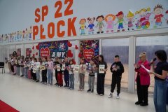  Na tle plakatów WOŚP dzieci z klas 1 – 3 ustawione w rzędzie prezentują dyplomy i pamiątkowe serca WOŚP. Są to nagrody za udział w aukcji prac plastycznych. 