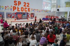 Zdjęcie przedstawia społeczność szkolną podczas uroczystego koncertu. Zebrani siedzą na podłodze i słuchają śpiewu wokalistów i chóru.  W tle widoczne są plakaty WOŚP. 