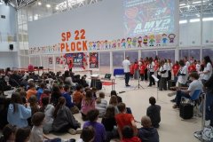 Na zdjęciu widoczny jest zespół muzyczny TuTu -  Band i chór TuTu. Artyści ubrani są w białe i czerwone koszulki z napisem TuTu. Dzieci siedzące na podłodze obserwują ich występ. 