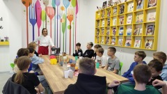 Grupa dzieci siedzi przy drewnianym stole i uczestniczy w warsztatach piernikowych prowadzonych przez kobietę w białym fartuchu i czerwonym fartuchu kuchennym. Na ścianie widać kolorowe widelce i łyżki jako dekorację oraz żółtą półkę z książkami kulinarnymi. Na stole znajdują się kolorowe kubki, miski i składniki potrzebne do pieczenia pierników.