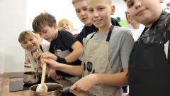 Grupa uczniów uczestniczy w warsztatach piernikowych, wspólnie mieszając składniki w garnkach i z uśmiechem pozując do zdjęcia. W tle widać kuchenne stanowisko pracy.