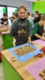 Dziewczynka w fartuchu z napisem "Cooking" stoi przy stole podczas warsztatów piernikowych, na macie silikonowej leżą wykrojone pierniki w różnych kształtach. W tle inni uczestnicy również pracują nad piernikami w kolorowej kuchni.