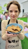 Dziewczynka w fartuchu z napisem "Cooking" trzyma miskę z upieczonymi piernikami podczas warsztatów piernikowych. W tle widać innych uczestników pracujących w kuchni z zielonymi szafkami i dekoracją w motyw liści.