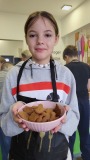 Dziewczynka w fartuchu z napisem "Cooking" trzyma miskę z upieczonymi piernikami podczas warsztatów piernikowych. W tle widać innych uczestników pracujących w kuchni z zielonymi szafkami i dekoracją w motyw liści.