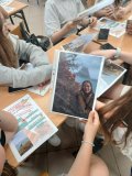 Zbliżenie na ławkę, przy której siedzą uczniowie. Na blacie leżą: używane podczas ćwiczeń smartfony, plakat z kolorowymi zasadami wykonywania interesujących zdjęć oraz kilka przykładowych, prawidłowo wykonanych zdjęć w plenerze.