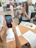 Zdjęcie grupki uczennic, gdzie dwie z nich wykonują sobie nawzajem zdjęcia smartfonami według instrukcji. W tle widać innych uczestników warsztatów- seniorów oraz prowadzącą.