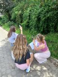 Ćwiczenia w plenerze- dwie uczennice i prowadząca- kucające wsród roślinności i wykonujące smartfonem ciekawe ujęcia.