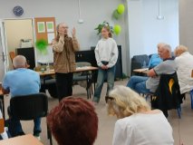 Zdjęcie zrobione z tyłu sali, w której zgromadzeni są seniorzy podczas warsztatów teatralnych, w dali widać reżysera współpracującego z przedstawicielką Samorządu Uczniowskiego.