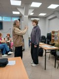 Reżyser podczas ćwiczeń z przedstawicielem SU, wokół drewniane ławki, przy których siedzą seniorzy.