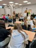 zdjęcie wykonane z tyłu sali, w której odbywają się warsztaty teatralne. W centrum znajduje się reżyser pracujący z przedstawicielką SU, a wokół przy drewnianych ławkach siedzą seniorzy i uczestnicy Koła SU.