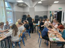 Zdjęcia przedstawiajace warsztaty plastyczne- w przestronnej sali przy ławkach ustawionych w literę "U" siedzą uczsetnicy: seniorzy i uczniowie z Koła SU, wszyscy w pogodnych nastrojach, współpracujący przy tworzeniu łapaczy promieni słońca. Na stoliczkach znajduje się mnóstwo elementów plastycznych: płyty CD, koraliki, cekiny, elementy odblaskowe itp.