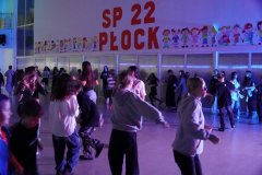 Uczniowie Szkoły Podstawowej nr 22 w Płocku bawią się podczas dyskoteki andrzejkowej na przestronnej sali, oświetlonej kolorowymi światłami. W tle napis „SP 22 Płock” oraz rysunki dzieci na ścianie.