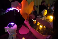 Uczniowie biorą udział w andrzejkowej wróżbie, trzymając duży papierowy kształt serca. Na stole widać zapalone sztuczne świece, dekoracje oraz kolorowe światła tworzące magiczny nastrój. 