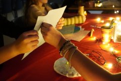 Uczniowie biorą udział w andrzejkowej wróżbie — jedna osoba trzyma kartę z wróżbami w kształcie serca, a druga przekłuwa ją w wybranym miejscu. Wokół migoczą kolorowe światełka, a na stole leżą tematyczne andrzejkowe rekwizyty i dekoracje. 