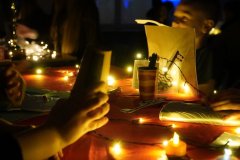 Zbliżenie na stół przykryty czerwonym obrusem, na którym znajdują się światełka, zapalone sztuczne świece oraz andrzejkowe rekwizyty, w tym ozdobny klucz. Ktoś trzyma kartę z wróżbami, a w oddali widać inne stanowiska wróżących uczniów. 