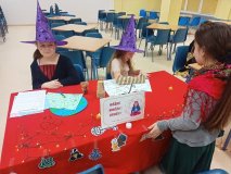 Dzieci w kapeluszach czarownic prowadzą wróżby przy czerwonym, ozdobionym lampkami stole. Na blacie znajdują się kartki z zadaniami, kolorowe rekwizyty oraz elementy dekoracji andrzejkowych. 