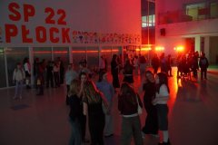  Uczniowie Szkoły Podstawowej nr 22 w Płocku bawią się podczas dyskoteki andrzejkowej na przestronnej sali, oświetlonej kolorowymi światłami. W tle napis „SP 22 Płock” oraz rysunki dzieci na ścianie.