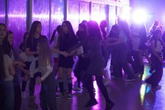 Uczniowie bawią się podczas dyskoteki andrzejkowej na przestronnej sali, oświetlonej kolorowymi światłami. 