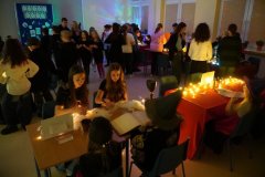 Uczniowie przebywają w andrzejkowej sali wróżb, siedząc przy udekorowanych stolikach ze świecami i rekwizytami. Część dzieci jest w przebraniach wróżek i czarodziejów, a w tle widać pozostałych uczestników wydarzenia. 