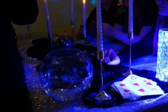 Zbliżenie na stół wróżb andrzejkowych, udekorowany świecami, kartami do wróżenia i szklaną kulą z losami. Wokół stołu siedzą uczniowie uczestniczący w zabawie.