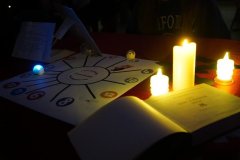 Zbliżenie na stół wróżb andrzejkowych, udekorowany sztucznymi świecami, planszą z wróżbami oraz otwartą księgą "Mały poradnik życia".