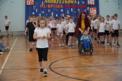 Dwoje dzieci z klas pierwszych, które wykonują zadanie polegające na przeniesieniu piłki tenisowej na drewnianej łyżce. Jedno z nich to chłopiec poruszający się na wózku inwalidzkim. W wykonaniu zadania pomaga mu nauczyciel. 