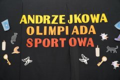 Na zdjęciu widać czarne tło z dużym kolorowym napisem: „Andrzejkowa Olimpiada Sportowa”. Wokół napisu znajdują się ozdobne ilustracje kojarzące się z andrzejkami i magią, m.in.: klucze, świece, kapelusz czarownicy, karty, koty, gwiazdki… Całość stanowi dekorację przygotowaną na szkolną imprezę andrzejkową o sportowym charakterze. 