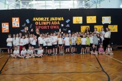 Na zdjęciu widać dużą grupę Dzieci oraz kilku Nauczycieli pozujących do wspólnego zdjęcia w sali gimnastycznej. W tle znajduje się czarna dekoracja z napisem „Andrzejkowa Olimpiada Sportowa” oraz kolorowe plakaty i rysunki przygotowane przez Uczniów. Uczestnicy stoją i siedzą w kilku rzędach, Dzieci mają stroje sportowe. Zdjęcie jest pamiątkową fotografią wszystkich Uczestników wydarzenia.